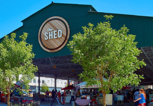 A large green building with "The Shed" sign, surrounded by trees and bustling market stalls.