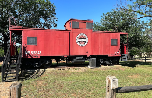 A red caboose on display in a grassy area, surrounded by trees and a clear blue sky.