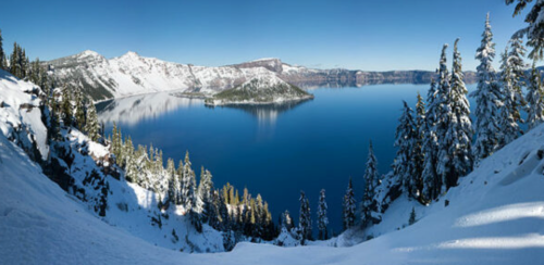 A panoramic view of a snowy landscape surrounding a calm, blue lake, with mountains in the background under a clear sky.