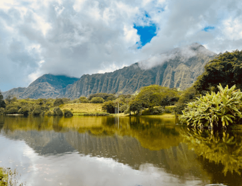 A serene lake reflects lush mountains and cloudy skies, surrounded by greenery and trees.