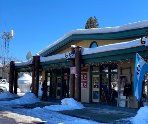 A snow-covered storefront with wooden beams, featuring signs for various shops and a clear blue sky above.