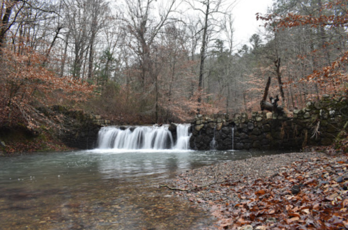 A serene waterfall cascades over a stone wall, surrounded by bare trees and autumn leaves along a calm river.