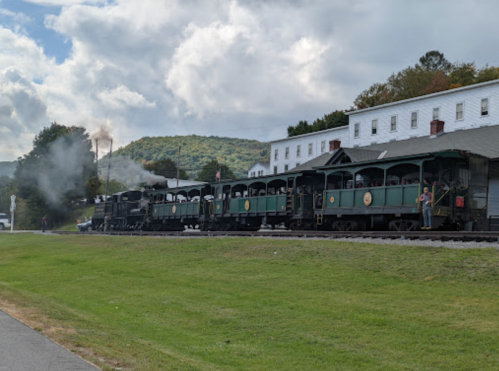 A vintage steam train with green carriages stops at a station, surrounded by hills and a cloudy sky.