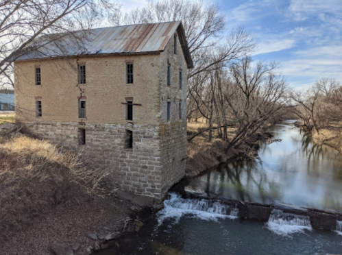 Abandoned stone building by a river, surrounded by bare trees and a small waterfall. Clear blue sky above.