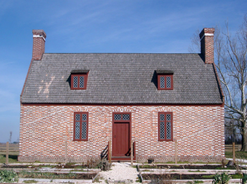 A historic brick house with a gray roof, featuring red windows and a central door, set against a clear blue sky.