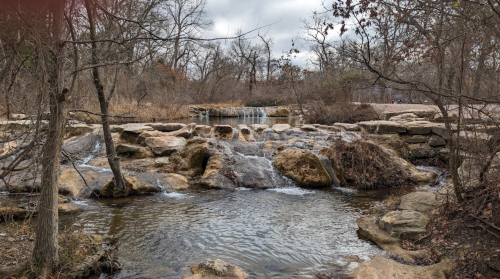 A serene landscape featuring a small waterfall cascading over rocks, surrounded by bare trees and a cloudy sky.