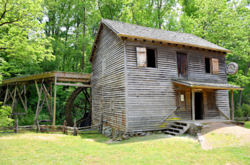 An old wooden mill with a water wheel, surrounded by lush green trees and grass.