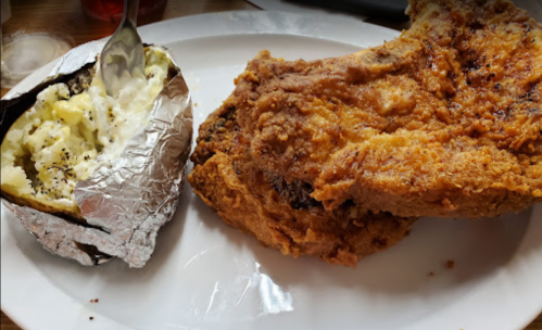 A plate featuring a large piece of fried chicken and a baked potato wrapped in foil.