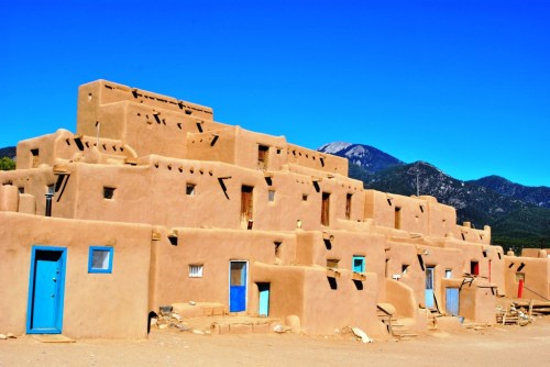 Adobe-style buildings with colorful doors against a clear blue sky and mountains in the background.