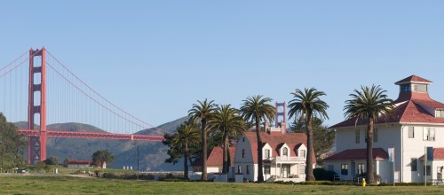 Golden Gate Bridge in the background with palm trees and historic buildings in the foreground. Clear blue sky.