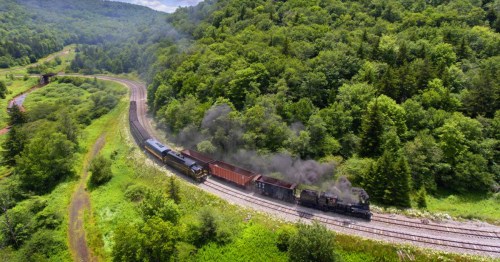 A train travels along a winding track through lush green forests, emitting smoke against a scenic landscape.