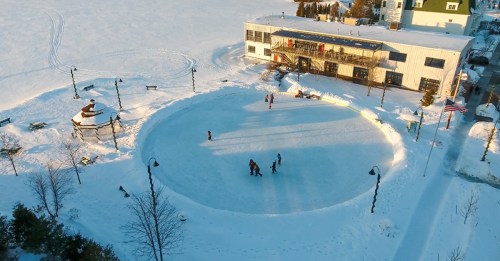 Aerial view of a circular ice skating rink surrounded by snow, with people skating and a building in the background.