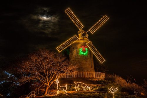 A lit windmill decorated for Christmas, surrounded by festive lights, under a moonlit sky.