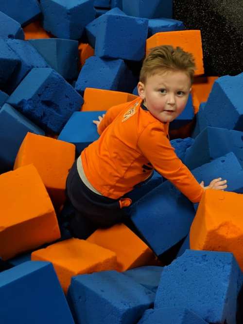 A young child in an orange shirt climbs through a pile of blue and orange foam blocks at a play area.