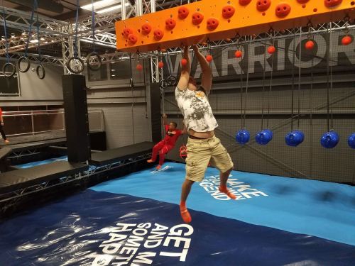 A person hangs from an orange climbing wall in an obstacle course gym, with another participant in the background.