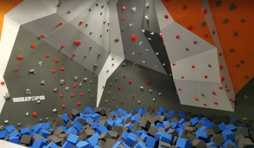 A climbing wall with colorful holds and a foam pit below, featuring gray, orange, and blue elements.