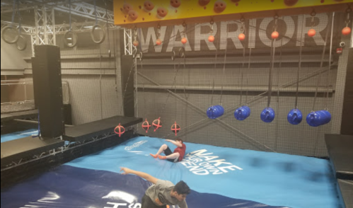 Two children play on a blue inflatable surface in a gym with climbing obstacles and a "WARRIOR" sign above.