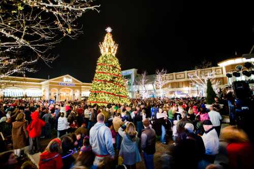 A large, decorated Christmas tree stands in a bustling plaza filled with people celebrating at night.