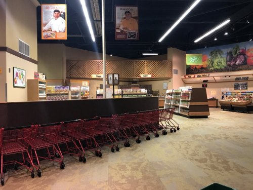 An empty grocery store aisle with shopping carts lined up and colorful produce displays in the background.