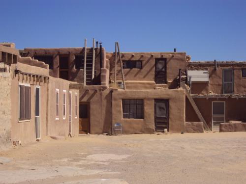 Adobe-style buildings with flat roofs and wooden ladders, set against a clear blue sky in a desert landscape.