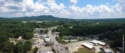 Aerial view of a small town surrounded by greenery and hills, with roads and buildings visible under a cloudy sky.