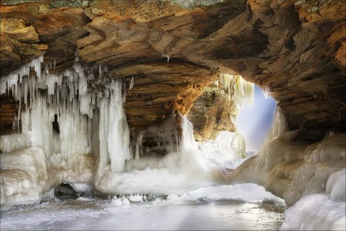 A frozen cave with icicles hanging from the ceiling and a bright light shining through an opening.