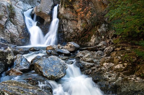 A serene waterfall cascades over rocks, surrounded by lush greenery and autumn foliage.