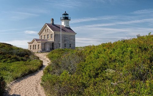 A historic lighthouse surrounded by greenery, with a sandy path leading to it under a clear blue sky.