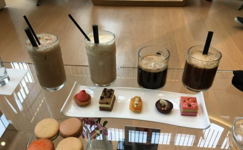 Four glasses of iced coffee drinks with straws, accompanied by a platter of assorted colorful desserts.