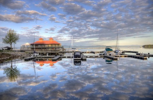 A serene lakeside scene with a dock, boats, and a building reflecting in calm water under a cloudy sky.