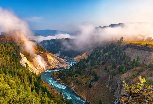 A winding river flows through a misty canyon, surrounded by lush trees and mountains under a clear blue sky.