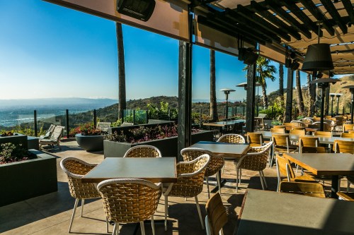 A scenic outdoor dining area with wicker chairs and tables, overlooking a valley and mountains under a clear blue sky.