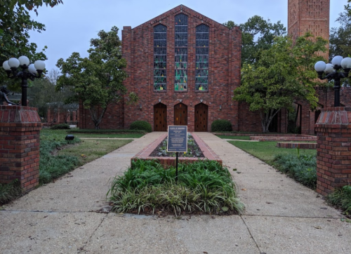 A brick building with large windows, flanked by trees and lamps, features a sign in front and a pathway leading to it.