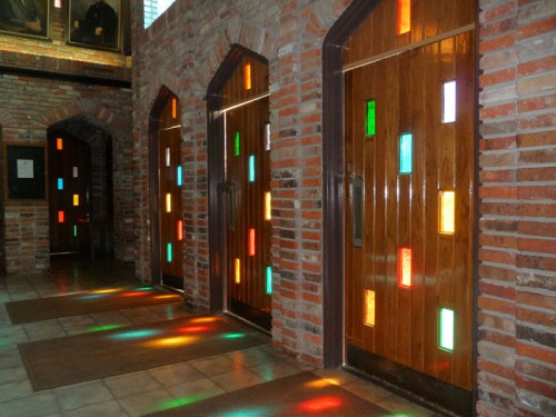 Three wooden doors with colorful glass panels, set in a brick wall, casting vibrant light patterns on the floor.