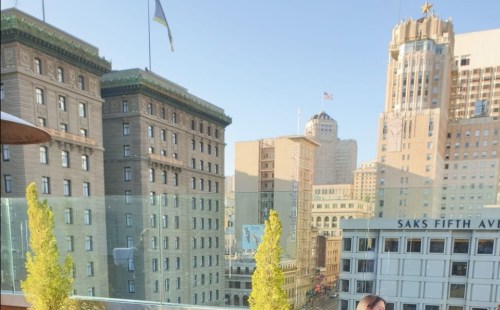 A cityscape featuring historic buildings, flags, and a clear blue sky, with a person in the foreground.