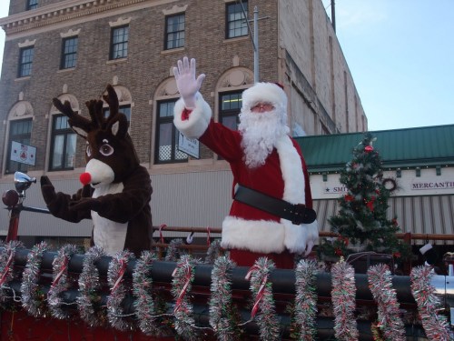 Santa Claus and a reindeer wave from a festive parade float, surrounded by holiday decorations and a Christmas tree.