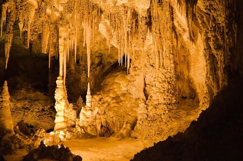 A dimly lit cave interior featuring impressive stalactites and stalagmites, illuminated by warm orange light.
