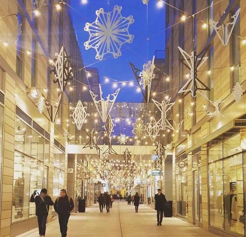A festive shopping alley decorated with snowflake ornaments and string lights, illuminated at dusk.