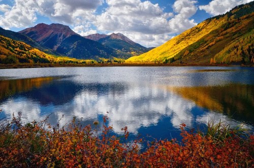 A serene lake reflects mountains and colorful autumn foliage under a bright, cloudy sky.