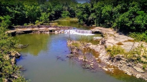 A serene river scene with a small waterfall surrounded by lush greenery and rocky banks.