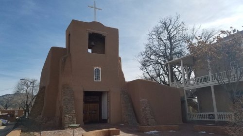 Adobe-style church with a cross on top, surrounded by trees and a clear blue sky.