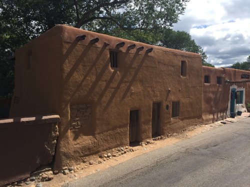 A rustic adobe building with a textured brown exterior, set against a cloudy sky and surrounded by greenery.