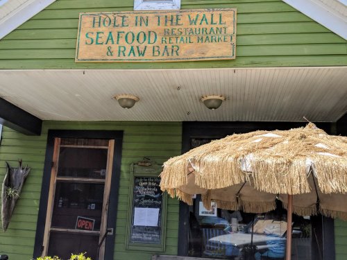 Sign for "Hole in the Wall Seafood Restaurant & Raw Bar" on a green building with a straw umbrella in front.