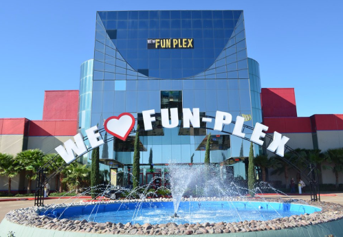 A modern building with a large sign reading "WE ❤️ FUN-PLEX" and a fountain in front, under a clear blue sky.