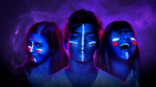 Three individuals with vibrant face paint in a colorful, smoky background, showcasing expressive emotions.
