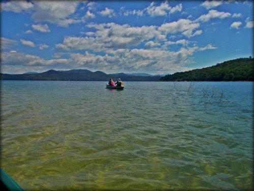 A small boat with people floats on a calm lake, surrounded by mountains and a blue sky with fluffy clouds.
