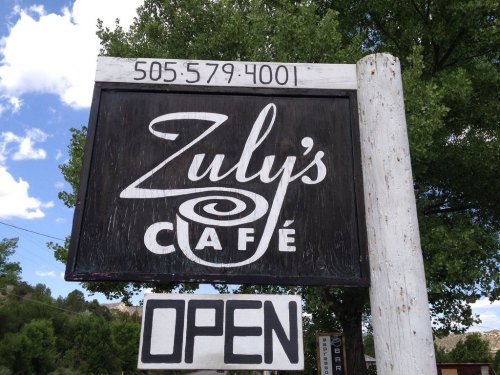 Sign for Zuly's Café with a phone number and "OPEN" displayed below, surrounded by trees and a blue sky.