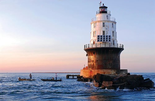 A lighthouse stands on a rocky shore at sunset, with two small boats nearby in calm waters.
