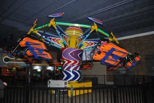 A colorful indoor amusement ride with spinning seats and bright lights, surrounded by a safety fence.