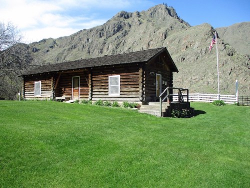 A rustic log cabin on green grass, with mountains in the background and an American flag nearby.